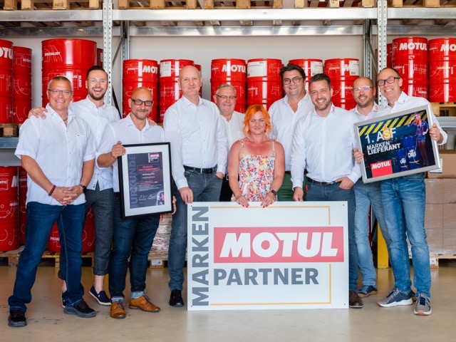 Das Team der ASF Jens Bauer oHG mit Urkunde und Schild zur MOTUL-Markenpartnerschaft vor roten MOTUL-Fässern im Lager.